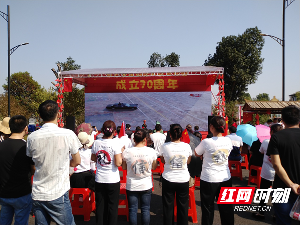 宁乡市关山古镇的电子大屏上，正播放着阅兵式，大伙儿纷纷围坐观看。