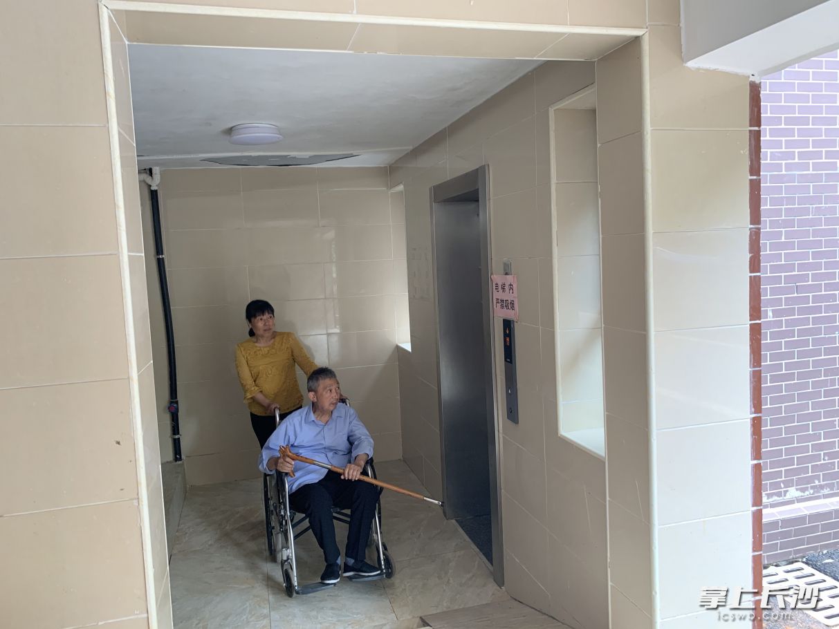 王文乐爹爹坐着轮椅，在保姆的帮助下，坐电梯到楼下逛公园。